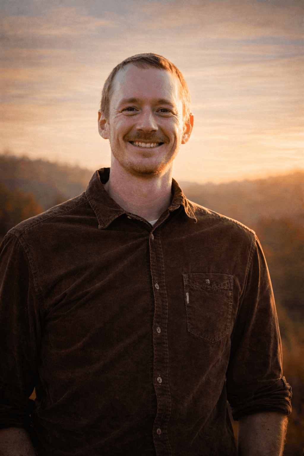 Smiling man with a mustache wearing a brown corduroy shirt during a golden sunset.