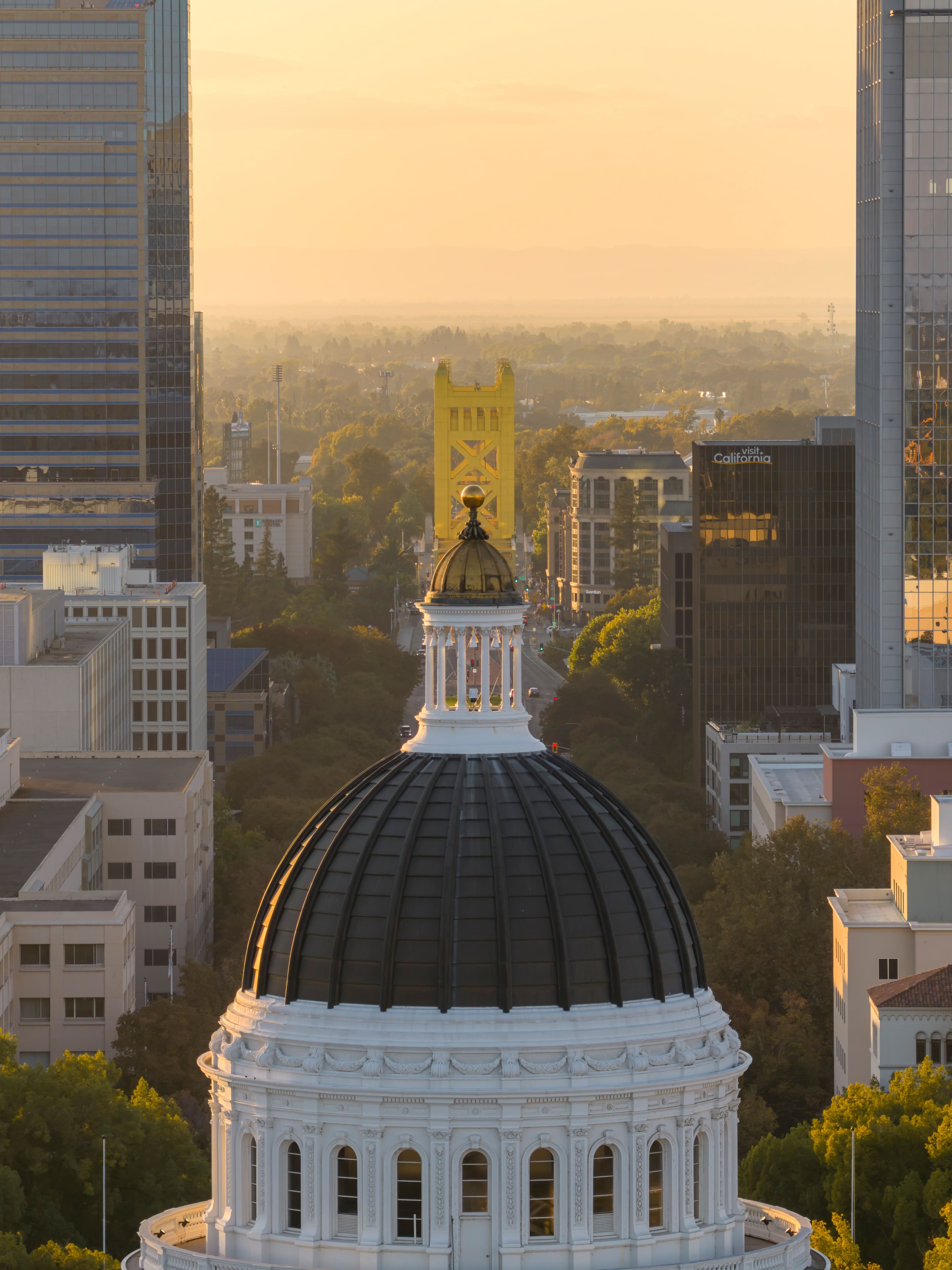 California State Capitol dome overlooks Sacramento's cityscape and the yellow Tower Bridge at sunset.